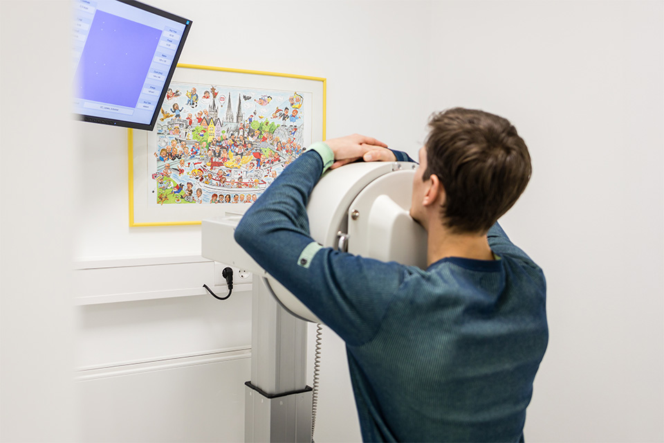 Mann sitzt mit dem Kopf vor einem Gerät für bildgebende Diagnostik, daneben Monitor und ein buntes Wimmelbild an der Wand.