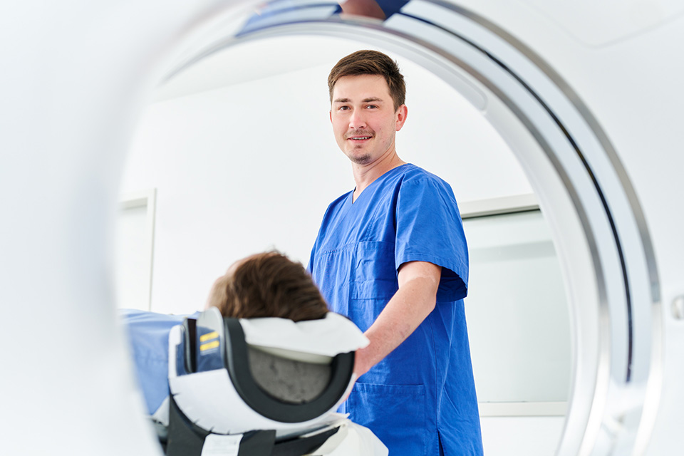 Medizinische Fachkraft in blauer Arbeitskleidung steht neben einer liegenden Person vor einem Gerät für bildgebende Diagnostik.