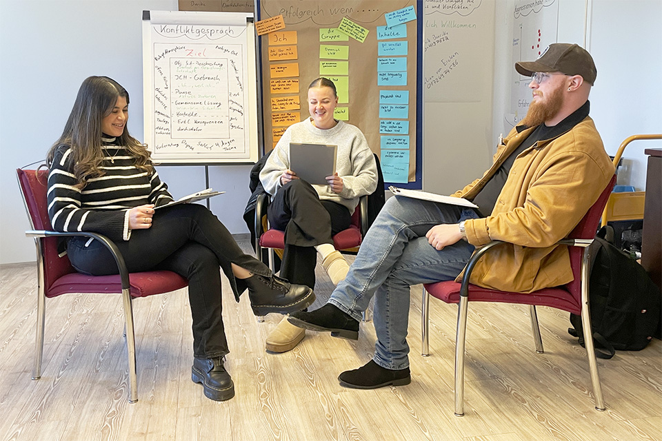 Kleine Gruppe in einem Seminarraum während einer interaktiven Übung; im Hintergrund sind ein Flipchart und eine Pinnwand mit erarbeiteten Workshop-Ergebnissen zu sehen.
