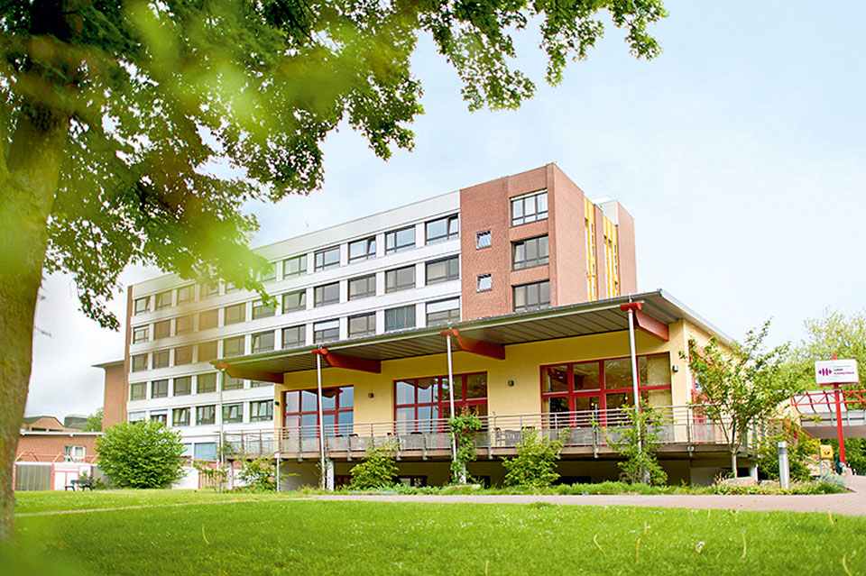 Das mehrstöckige Lukas-Krankenhaus in Gronau mit rotem und weißem Fassadenteil, davor ein niedrigerer gelber Anbau mit überdachtem Eingang und Grünfläche im Vordergrund